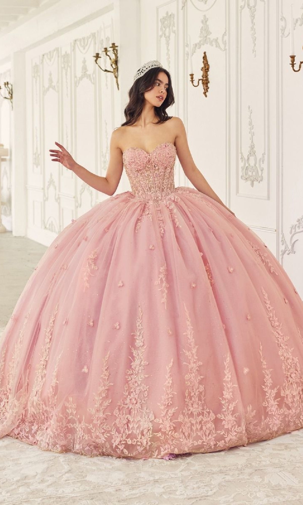 A young woman in the Quinceanera Dress 15723 by Ladivine, featuring a strapless design and floral lace applique, stands in an ornate white room with elegant wall detailing, wearing a tiara.